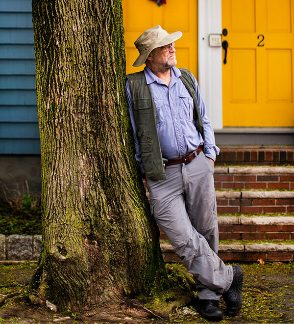Lawrence Millman wearing a hat and khaki vest leans against a tree in front of a yellow door