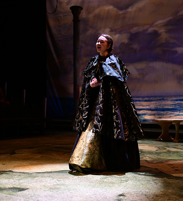 Ella Sherlock plays Hermione, wearing a cloak in spotlight on stage
